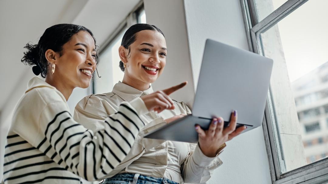 Kaksi hymyilevää henkilöä katsoo tietokonetta. Tautalla näkyy vaalea seinä ja ikkunasta kaupunkimaisema. 