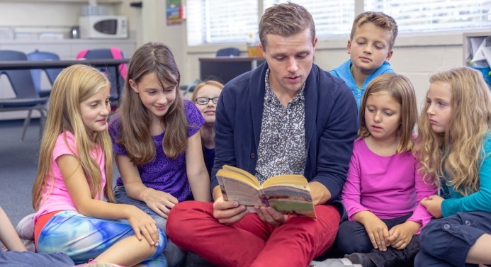 Lapsia varhaiskasvatuksen ammattilaisen kanssa lukemassa satua. Taustalla päiväkodin luokkahuone.